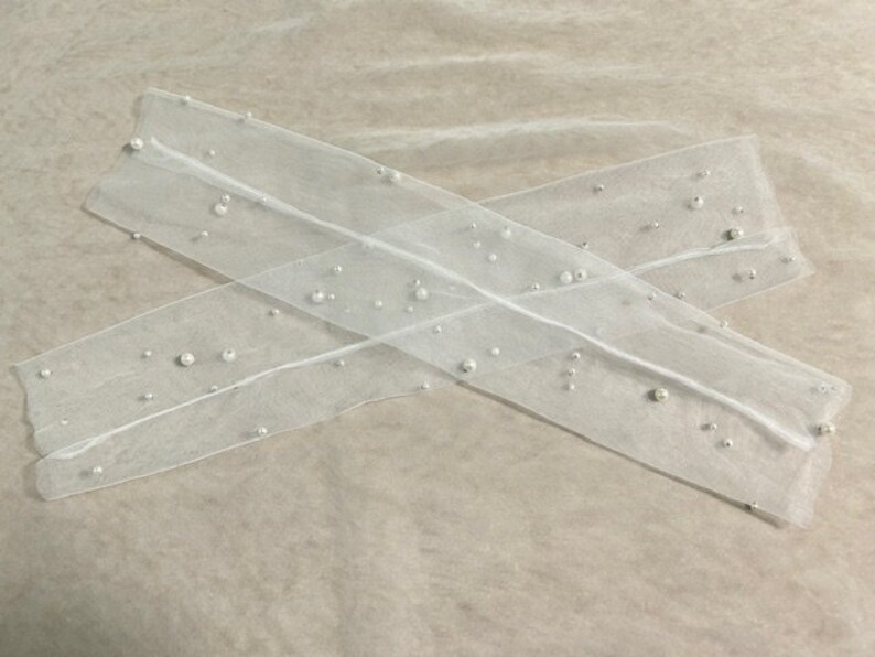 Pu&ograve; includere: Due strisce di tessuto bianco trasparente, ciascuna adornata con perle finte sparse di varie dimensioni. Le strisce sono disposte a croce, creando un'estetica delicata ed elegante. Lo sfondo &egrave; beige chiaro neutro.