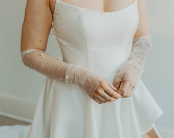 May include: A white bridal gown with a fitted bodice and spaghetti straps. The dress features a flared skirt and is paired with sheer, long gloves adorned with small, scattered pearls. The overall look is elegant and suitable for a wedding.