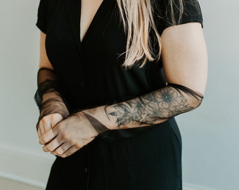 Può includere: Una donna indossa un abito nero con bottoni sul davanti e guanti trasparenti neri. I guanti hanno un motivo floreale sul dorso della mano.