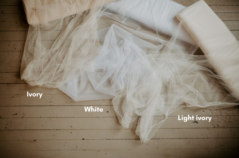 Pu&ograve; includere: Un assortimento di tessuti in tulle nei colori avorio, bianco e avorio chiaro. Il tulle &egrave; drappeggiato e arrotolato, mostrando la sua delicata consistenza e le variazioni di colore. L'immagine &egrave; ambientata su uno sfondo di legno rustico.