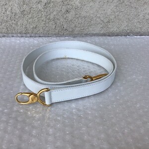 May include: A white leather purse strap with gold-colored metal clasps. The strap is coiled and lies on a textured surface. The clasps have a decorative design.
