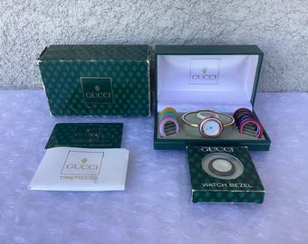 May include: A vintage Gucci watch set with a gold-tone bracelet, a white and red watch face, and a variety of colored bezels. The set includes the original box, a watch bezel box, and paperwork. The Gucci logo is visible on the box and paperwork.