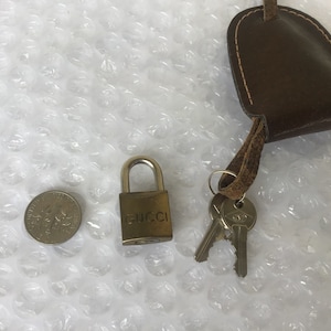May include: A small, gold-colored padlock with the word "GUCCI" engraved on the front. The padlock is attached to a brown leather strap with two keys.