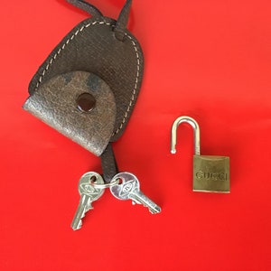 May include: A brown leather key holder with a metal key ring and two keys. A gold padlock with the word "GUCCI" engraved on it is also pictured.
