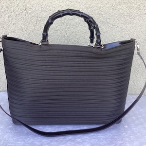 May include: Black tote bag with a bamboo handle and a detachable shoulder strap. The bag has a ribbed texture and silver-tone hardware. The handle is curved and glossy. The bag is designed with a zipper closure.