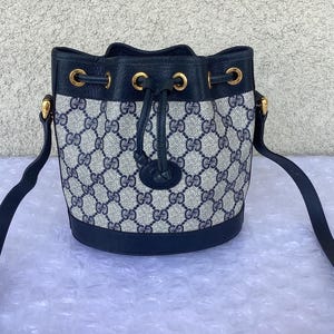 May include: A black and blue Gucci bucket bag with a gold chain strap and a drawstring closure. The bag is made of a canvas material with a GG pattern.