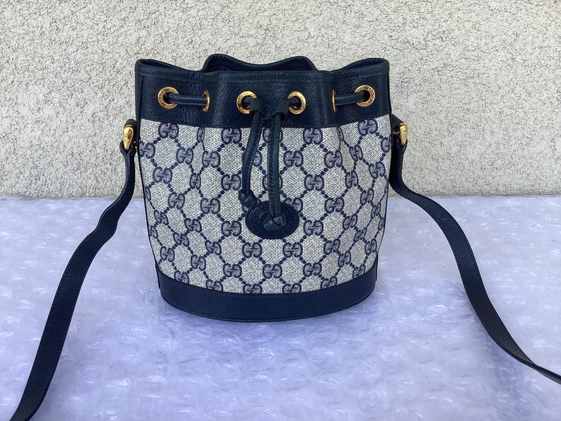 May include: A black and blue Gucci bucket bag with a gold chain strap and a drawstring closure. The bag is made of a canvas material with a GG pattern.