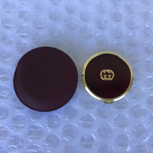 May include: A gold compact mirror with a red interior and the Gucci logo. The mirror comes with a burgundy leather case.