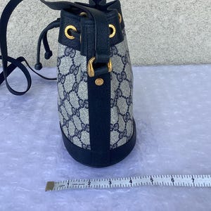 May include: A blue and beige Gucci bucket bag with a black leather trim and gold hardware. The bag has a drawstring closure and a shoulder strap.