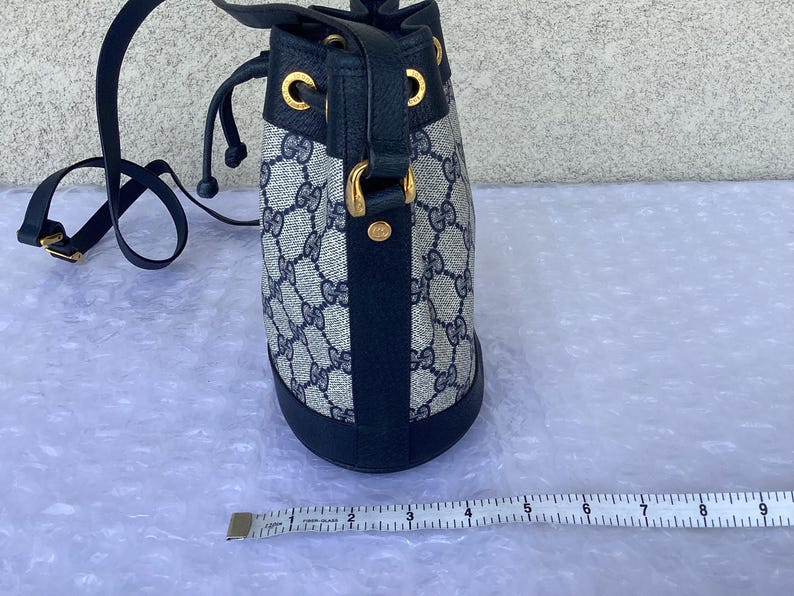 May include: A blue and beige Gucci bucket bag with a black leather trim and gold hardware. The bag has a drawstring closure and a shoulder strap.
