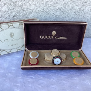 May include: A vintage Gucci Accessory Collection watch set in a brown velvet-lined box. The set includes a gold-toned watch with a white face and colorful interchangeable bezels. The box and packaging feature the Gucci logo.