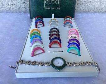 May include: A Gucci Timepieces watch set featuring a silver-tone metal link bracelet and a round watch face with a green and red bezel. The set includes a collection of interchangeable colorful circular bezels arranged in a white display tray.