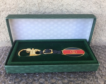 May include: A gold-colored key chain with a black leather strap and a gold oval charm with a red, black, and gold Gucci logo. The key chain is in a green velvet box.