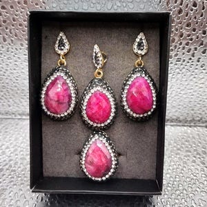 May include: A jewelry set consisting of two teardrop-shaped earrings and a matching pendant, each featuring a faceted hot pink stone surrounded by a border of small clear crystals set in a dark metal frame, presented in a black display box.
