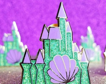 May include: A green and purple glitter castle pin with a purple seashell in the center. The pin is on a sparkly gold background.
