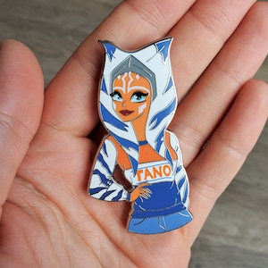 May include: A white and blue enamel pin featuring a cartoon character with blue eyes, orange skin, and white and blue striped hair. The character is wearing a blue and white striped shirt with the word "TANO" printed on it.