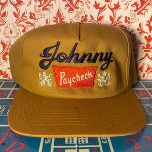 Puede incluir: Gorra de b&eacute;isbol color mostaza con visera plana. La gorra presenta el texto bordado "Johnny Paycheck" en azul marino y rojo, con emblemas decorativos a ambos lados del texto.