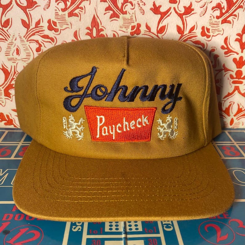 Puede incluir: Gorra de b&eacute;isbol color mostaza con visera plana. La gorra presenta el texto bordado "Johnny Paycheck" en azul marino y rojo, con emblemas decorativos a ambos lados del texto.