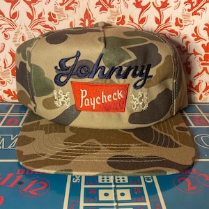 Puede incluir: Gorra de b&eacute;isbol con estampado de camuflaje con el nombre "Johnny" bordado en escritura azul marino sobre un rect&aacute;ngulo rojo con la palabra "Paycheck". La gorra tiene una visera plana y un cierre trasero cl&aacute;sico.