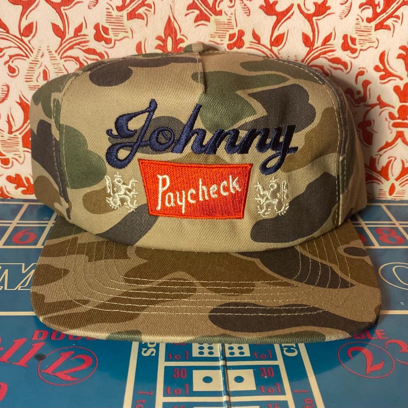 Puede incluir: Gorra de b&eacute;isbol con estampado de camuflaje con el nombre "Johnny" bordado en escritura azul marino sobre un rect&aacute;ngulo rojo con la palabra "Paycheck". La gorra tiene una visera plana y un cierre trasero cl&aacute;sico.