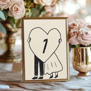 Puede incluir: Un número de mesa enmarcado en dorado con una ilustración en blanco y negro de una pareja sosteniendo un corazón con el número 1. El fondo es crema. La imagen está sobre una mesa con arreglos florales.