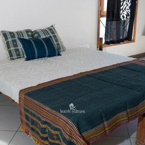 May include: A white bed with a dark blue and green throw blanket with a geometric pattern. The bed has a wooden frame and is decorated with pillows in shades of green and blue.