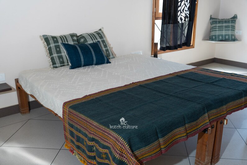 May include: A white bed with a dark blue and green throw blanket with a geometric pattern. The bed has a wooden frame and is decorated with pillows in shades of green and blue.