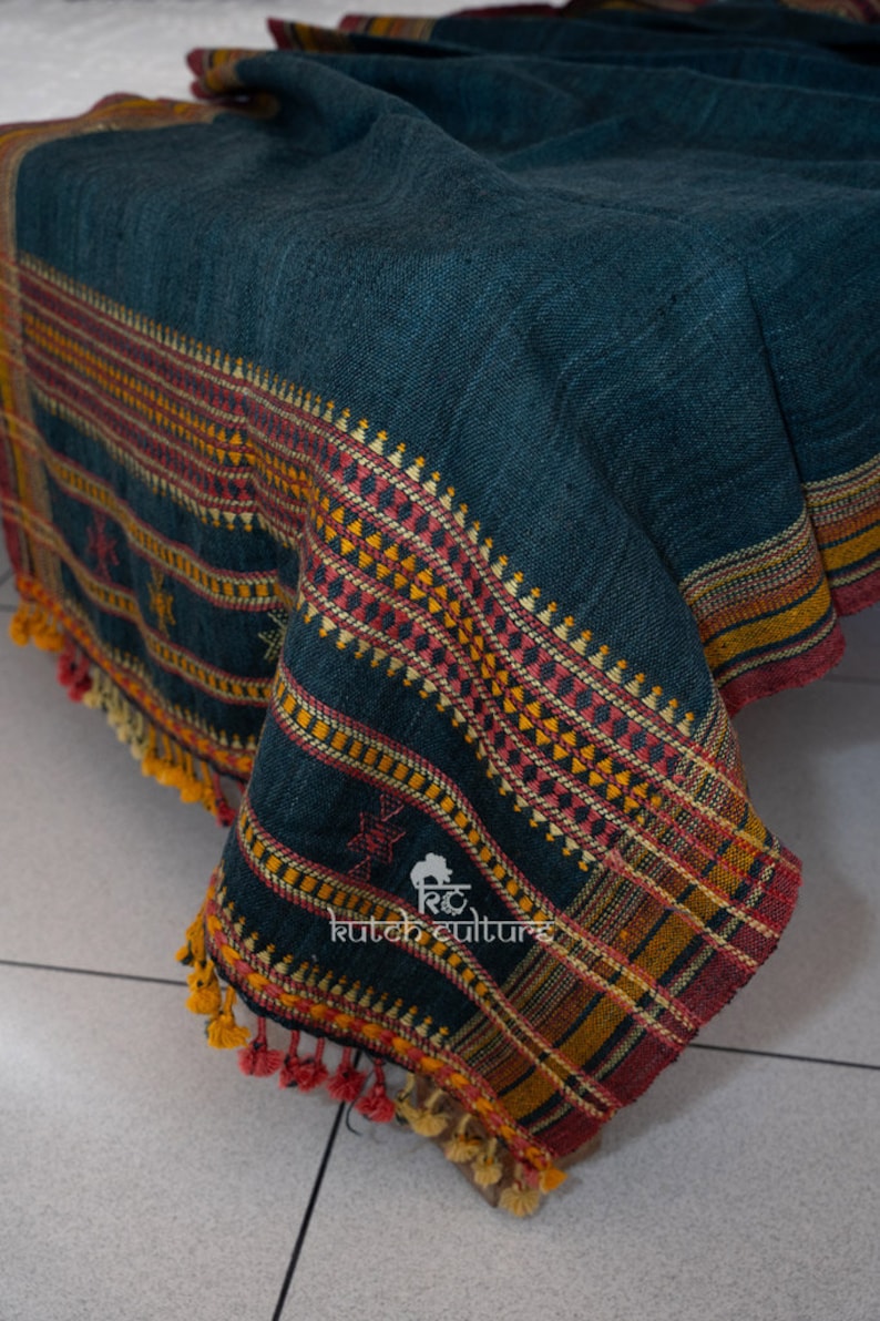 May include: A dark teal blue woven throw blanket with a colorful geometric pattern border. The blanket has a fringe edge and is made of wool.