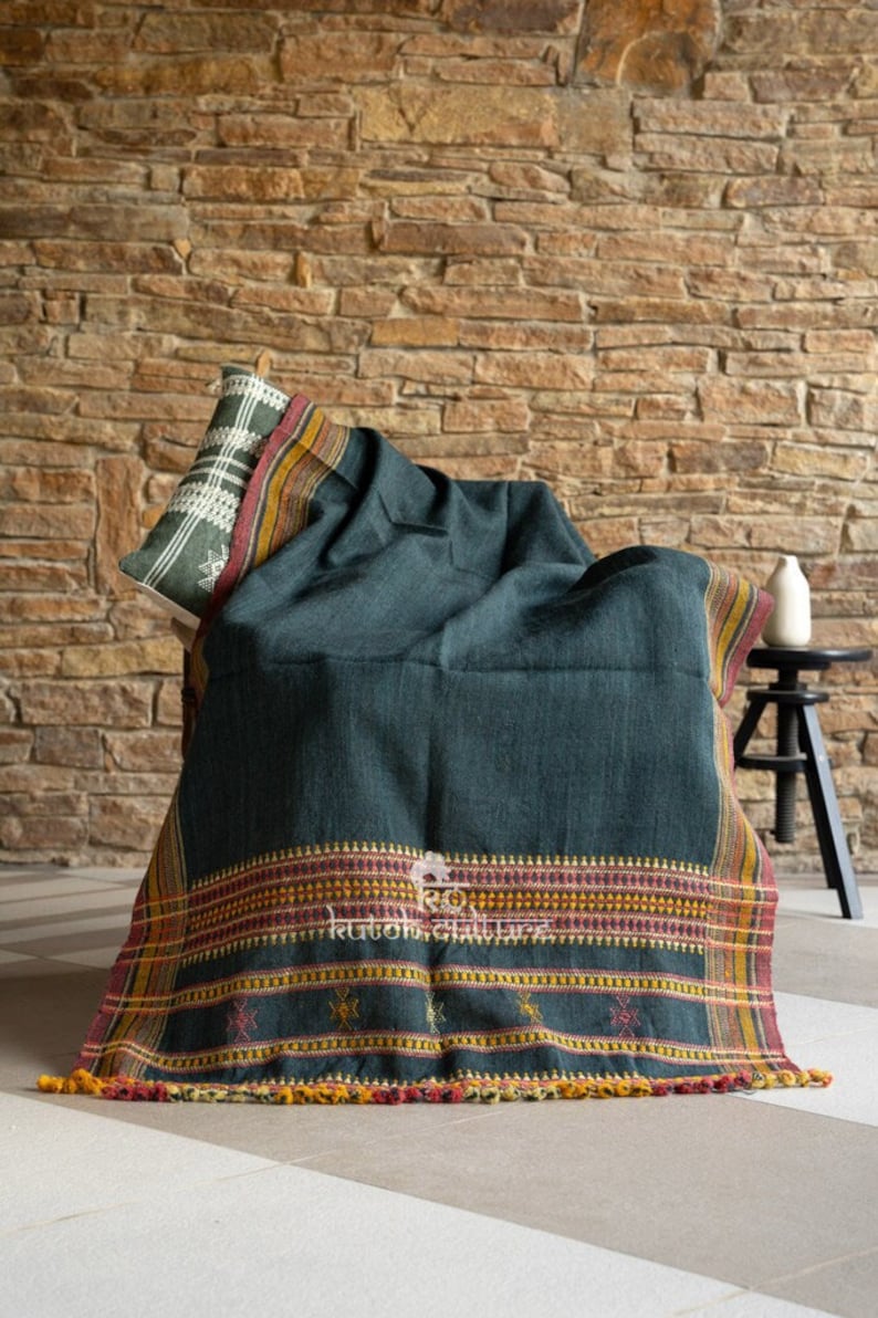 May include: A dark green throw blanket with a colorful woven border and tassels. The blanket is draped over a chair in front of a brick wall.