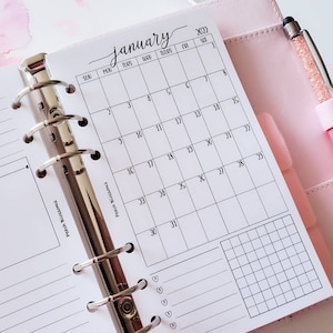 May include: A white monthly planner with a silver binder. The planner is open to January 2022 and has a grid layout with days of the week labelled. The planner has a section for notes and a grid for tracking habits.