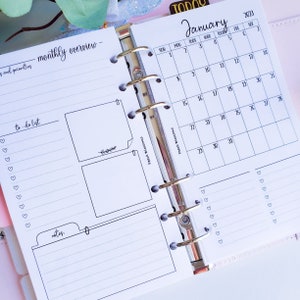 May include: A white planner with a monthly calendar for January 2023. The calendar has a to-do list, notes section, and space for monthly goals. The planner has a silver binder.