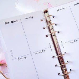 May include: A white planner with a silver binder. The planner has a weekly layout with the days of the week labeled: Monday, Tuesday, Wednesday, Thursday, Friday, Saturday. The planner also has the words "This Week" at the top.