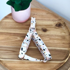 May include: A white dog harness with a pink, gold and grey terrazzo pattern. The harness has gold metal hardware.