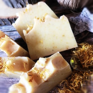 May include: Close-up of four bars of handmade soap with a pale yellow colour and flecks of dried flowers. The soap is sitting on a wooden surface.