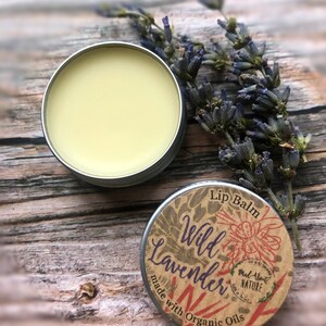 May include: A small tin of lip balm with a label that reads "Lip Balm Wild Lavender Made About Nature" and "made with organic oils". The tin is sitting on a wooden surface with dried lavender flowers.