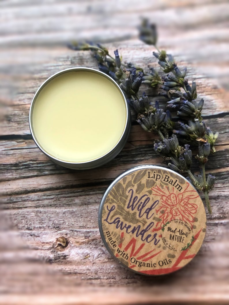 May include: A small tin of lip balm with a label that reads "Lip Balm Wild Lavender Made About Nature" and "made with organic oils". The tin is sitting on a wooden surface with dried lavender flowers.