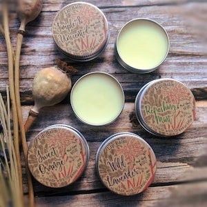 May include: Five small round metal tins of lip balm with brown paper labels. The labels have illustrations of flowers and the text "Lip Balm" and different flavors: "Simple & Unscented", "Sweet Orange", "Wild Lavender", "Refreshing Mint".