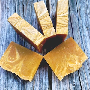 May include: Five bars of handmade soap with a swirl pattern in shades of yellow and white. The soap is arranged in a star shape on a wooden surface.
