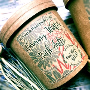 May include: A brown paper container with a label that reads "Natural & Handmade Soap Warming Winter Bath Salts". The label has a floral design with red and grey accents. The container is labelled "Mad About Nature" and has a website address.