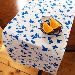 May include: A white table runner featuring a repeating blue floral and vine pattern. Resting on the runner is a small, white, scalloped-edge ceramic bowl decorated with blue floral motifs, containing two fresh orange slices.