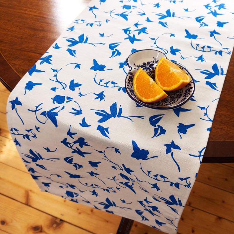 May include: A white table runner featuring a repeating blue floral and vine pattern. Resting on the runner is a small, white, scalloped-edge ceramic bowl decorated with blue floral motifs, containing two fresh orange slices.