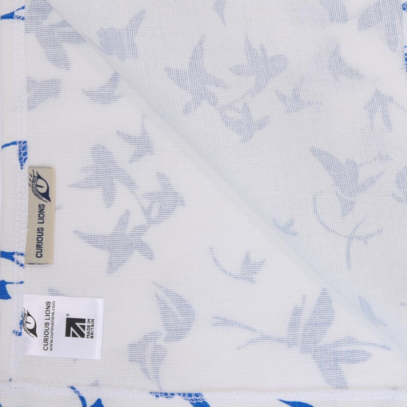May include: A white textile featuring a repeating pattern of blue bird silhouettes. The fabric includes a beige tag with the text CURIOUS LIONS and a white rectangular label displaying the text CURIOUS LIONS, curiouslions.com, and MADE IN BRITAIN.