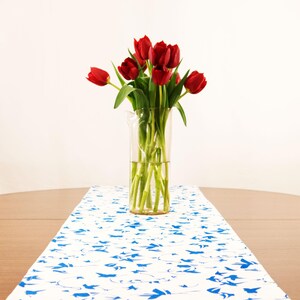 May include: A clear glass pitcher filled with a bouquet of vibrant red tulips with green stems and leaves. The pitcher sits on a white table runner featuring a repeating pattern of small blue birds in flight across the fabric surface.