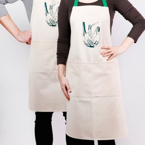 May include: Two white aprons with green straps and a black and white graphic of a fennel bulb on the front. The aprons have two pockets.