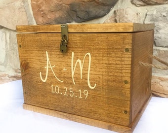Peut inclure: Coffret souvenir en bois avec couvercle à charnière et fermoir en métal. La boîte est teinte en brun chaud et présente les initiales "A + M" et la date "10.25.19" en écriture cursive. Une poignée en corde est attachée sur le côté.