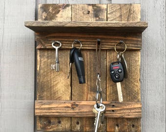 May include: A rustic wooden key holder with a small shelf. The holder is made of reclaimed wood and features several hooks for keys. Various keys, including car keys, are hanging from the hooks. The wood has a natural, weathered appearance.