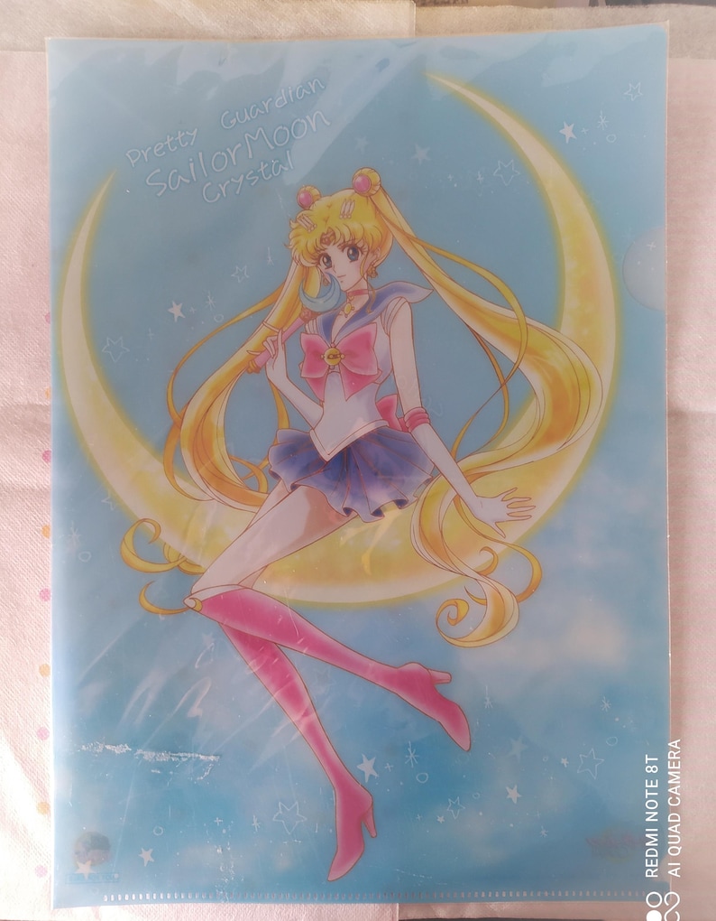 K&ouml;nnte beinhalten: Ein durchsichtiger Plastikordner mit einer bunten Illustration von Sailor Moon, die auf einem Halbmond steht. Der Text "Pretty Guardian Sailor Moon Crystal!" ist auf dem Ordner aufgedruckt.