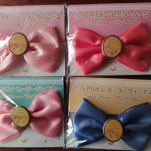 May include: Four pink, red, and blue hair bows featuring Sailor Moon and My Melody characters. Each bow has a different character and is labelled with the character's name.
