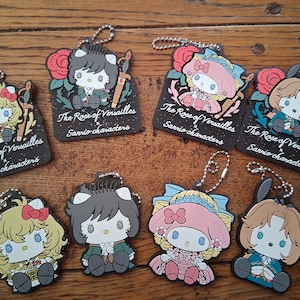Può includere: Collezione di portachiavi con personaggi Sanrio con disegni de "La Rosa di Versailles". Ogni portachiavi presenta un'illustrazione unica del personaggio con un design a rosa e spada. Il testo "The Rose of Versailles Sanrio characters" è stampato su ogni portachiavi.