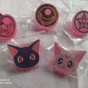 Può includere: Cinque ciondoli in plastica rosa e bianca con personaggi dell'anime Sailor Moon. I ciondoli hanno la forma di un cuore, una stella, una luna crescente e due facce di gatto.